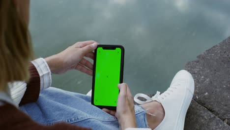 mujer sosteniendo un teléfono inteligente con pantalla verde al aire libre por el agua