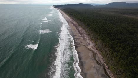 aerial view of forested coastline with ocean waves splashing on sandy shore. maori beach in bruce bay, new zealand.