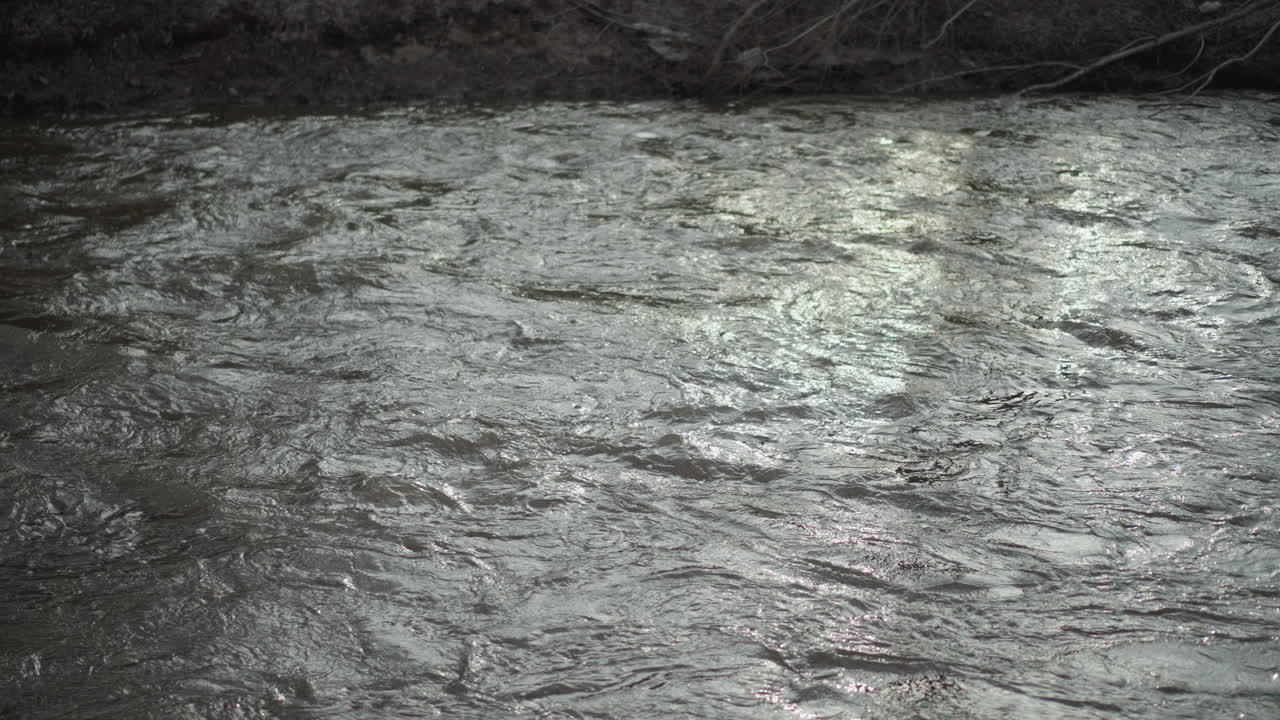 Closeup view of flowing river water surface with shimmering reflections under natural daylight, gentle ripples and small waves moving downstream, creating textured pattern and dynamic movement