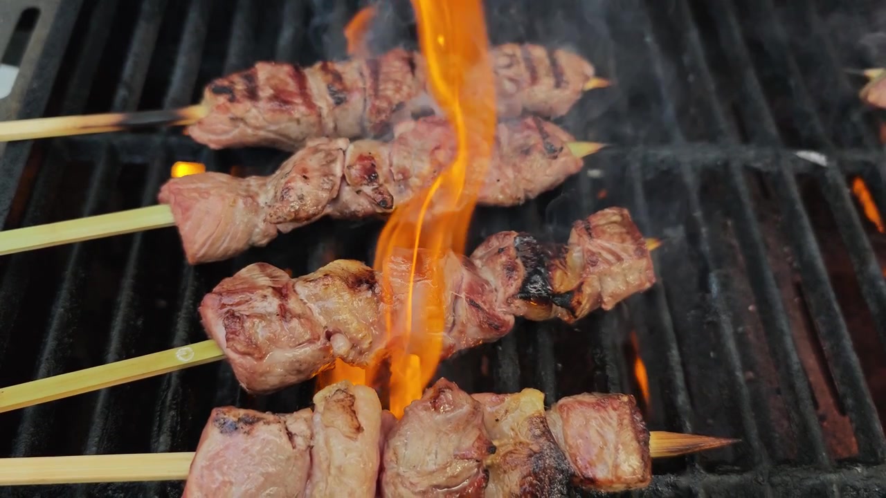 Video orizzontale in slow motion 4K di spiedini di manzo in preparazione su una griglia per barbecue, con fiamme, fumo e carne che sfrigola, che cattura la cucina all'aperto, la grigliata e la preparazione di cibo delizioso