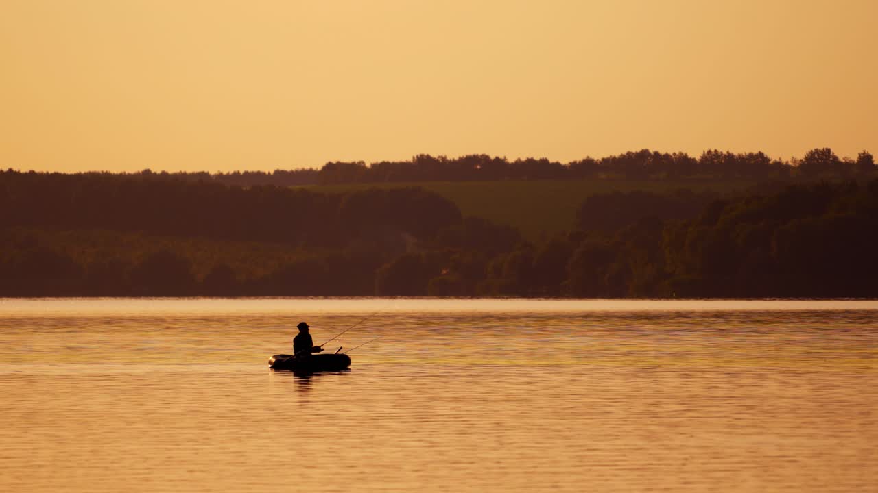Silhouette of fisherman in a boat on the natural rural evening background. Beautiful sunset on the river and a man in a rowboat with fishing rods outdoors.