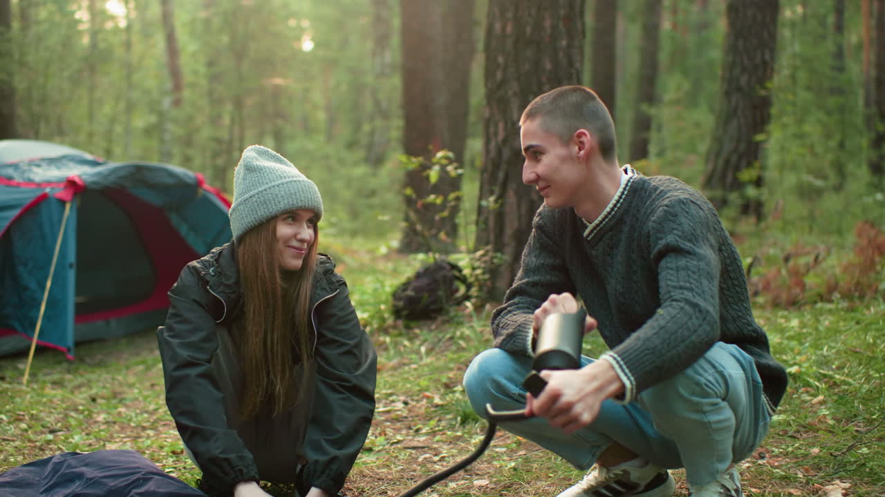 Couples sharing warm smiles while man pumps air into tent bag during camping activity, expressing connection and joy in peaceful outdoor setting with relaxed body