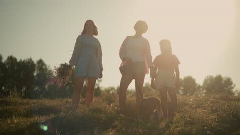 family standing on hilly farmland during sunset with sun shining behind them as mother reach out for her daughter hand, featuring two women and child with dog