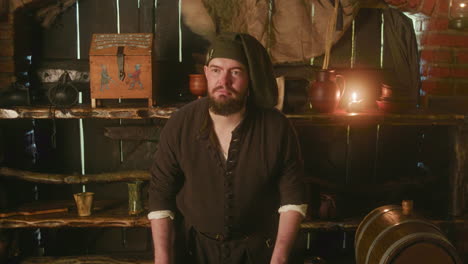 bearded keeper in bonnet waits for guests in tavern. stubby dwarf innkeeper stands by counter against rustic wooden shelves in store. fantasy character