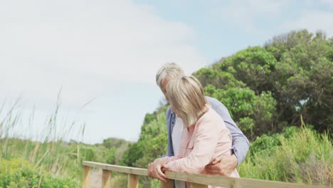 smiling senior caucasian couple embracing and standing on bridge outdoors
