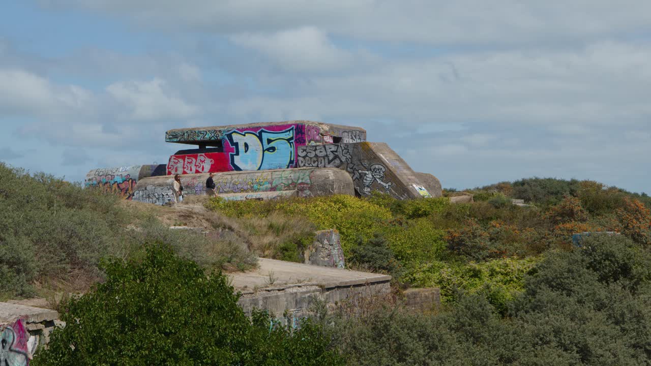 Drone pans across abandoned concrete fortification, graffiti art, overgrown vegetation, bright daylight, coastal landscape