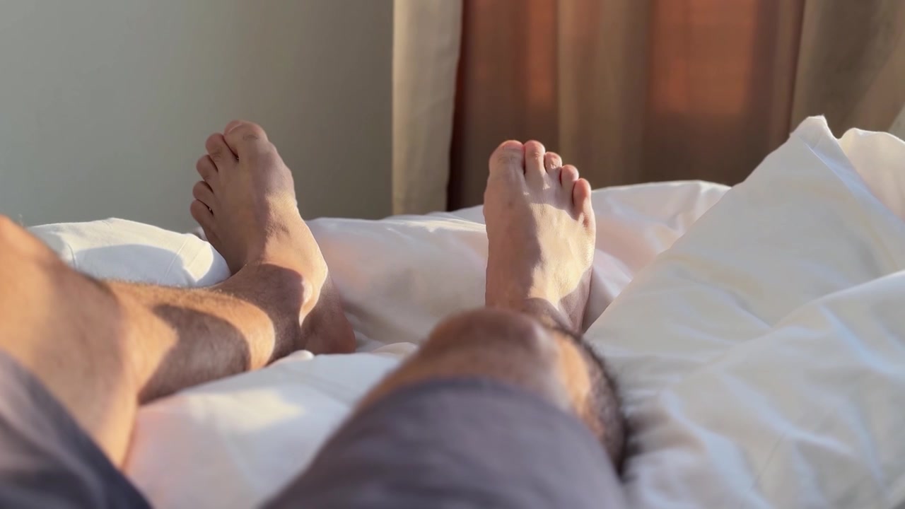 Bare male legs wearing shorts rest comfortably on white bed sheets in a bright indoor bedroom. Warm morning sunlight shines through brown window curtains, highlighting a relaxing vacation.