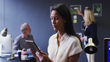 Mixed-race-businesswoman-using-digital-tablet-in-office-with-colleagues-discussing-behind