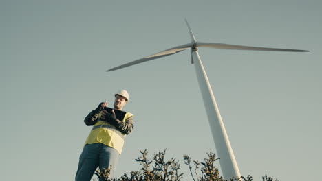 advancing renewable energy, an engineer uses a tablet to audit wind turbines in a field of clean energy generators on a sunny day, wearing a white helmet and reflective vest