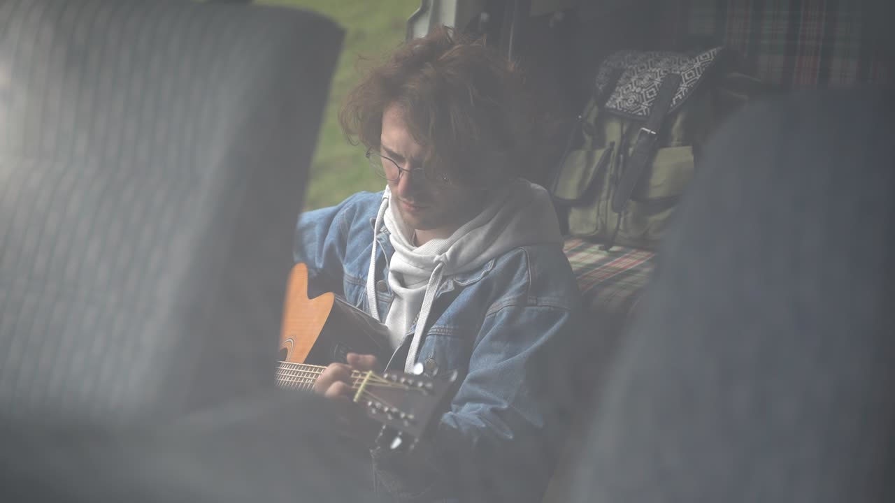 안경을 쓴 젊은 남자가 캐러밴 뒤에서 기타를 연주합니다.