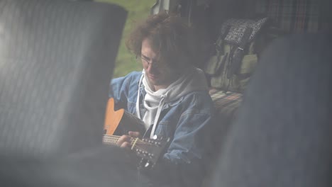 a young guy with glasses plays the guitar in the back of a caravan.