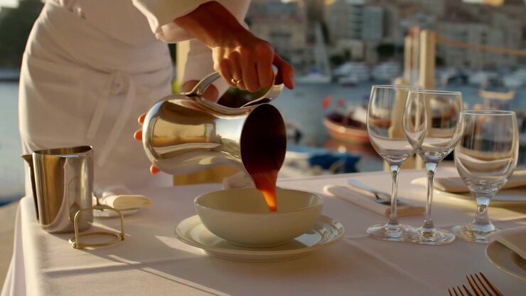 Warm low-angle sunlight crosses a neatly set terrace table as hands tilt a reflective pitcher, filling a shallow white bowl while clear glasses and moored boats remain softly blurred behind.