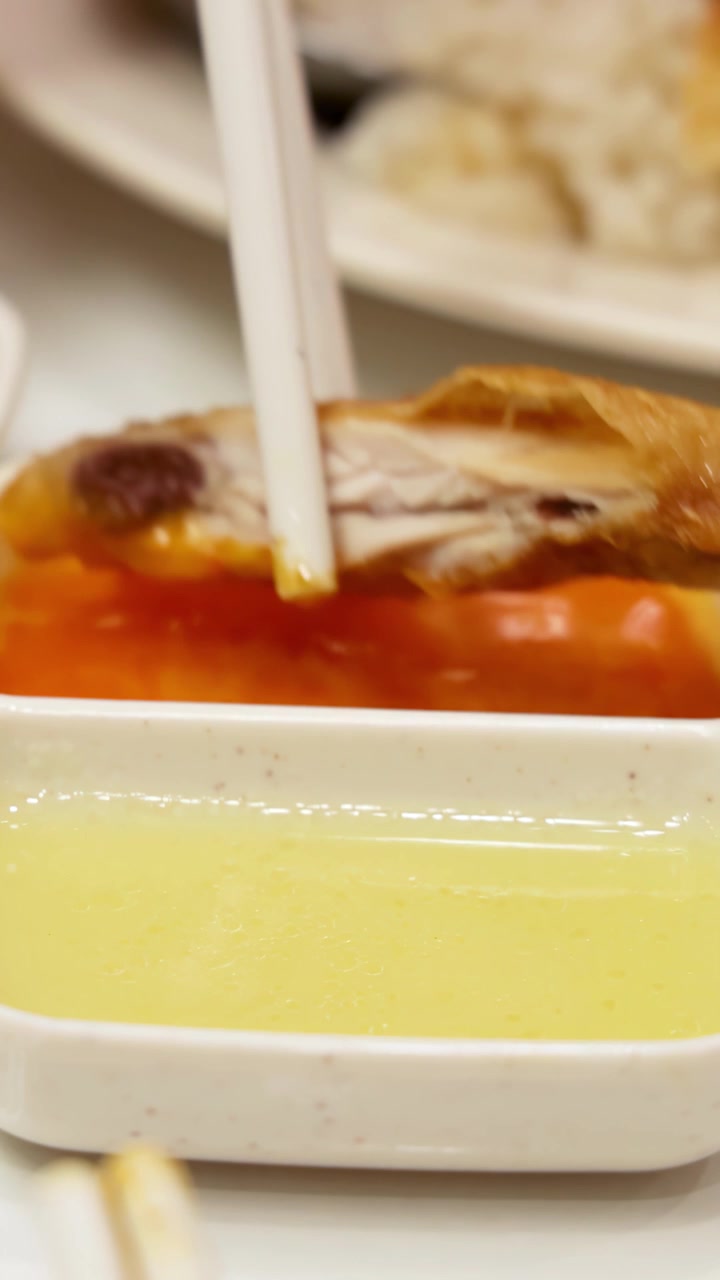 Il pollo arrosto viene preso con le bacchette e intinto in una vivace salsa chili, con riso Hainanese e utensili sullo sfondo illuminato delicatamente