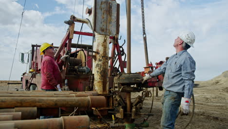 two men in protective helmets stand at the truck with drilling equipment. long metal pipe is pressed into the ground and water comes off.