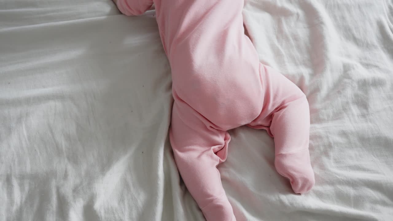 Neonato con vestitino rosa che tenta di gattonare su un letto bianco, vista dall'alto.