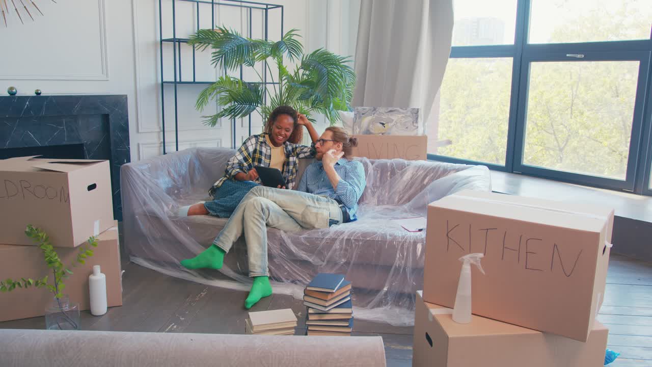 다민족 디자이너 부부가 리노베이션 계획을 논의하면서 상자 사이 소파에 앉아 있습니다.