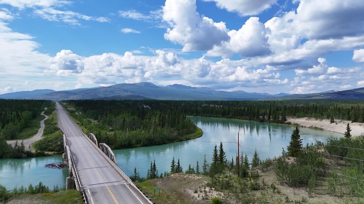 Un vasto ponte autostradale dell'Alaska attraversa un vivace fiume turchese, circondato da una fitta foresta sempreverde che conduce a montagne maestose sotto un cielo nuvoloso spettacolare
