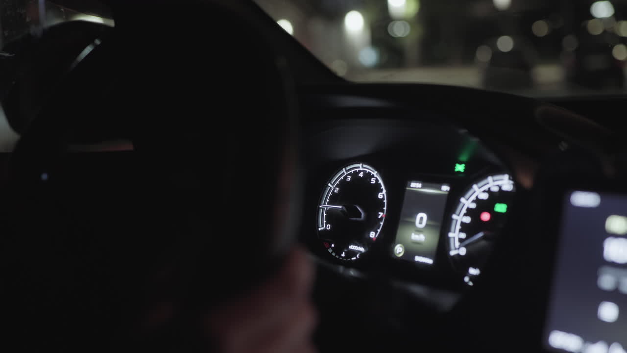 Detailed close up of illuminated modern car dashboard interior showing speedometer and indicator lights at night with blurred lights of passing cars visible on street through windshield