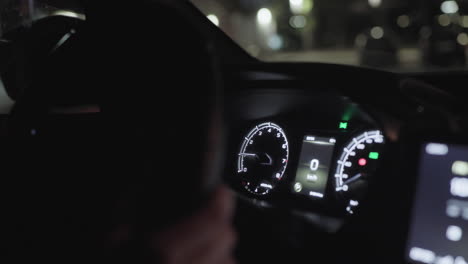 Detailed close up of illuminated modern car dashboard interior showing speedometer and indicator lights at night with blurred lights of passing cars visible on street through windshield