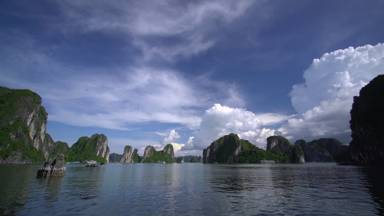 เรือแล่นช้าๆ ผ่านผืนน้ำที่สงบ ล้อมรอบด้วยเกาะหินปูน ใต้ท้องฟ้าสีครามพร้อมเมฆปุยคล้ายปุยฝ้ายในอ่าวฮาลอง