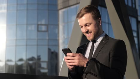 happy businessman holding a smartphone and laughing happy businessman holding a smartphone and laughing joyful manwinner with a gadget in his hands a man in business clothes stands near a skyscraper and reads the news