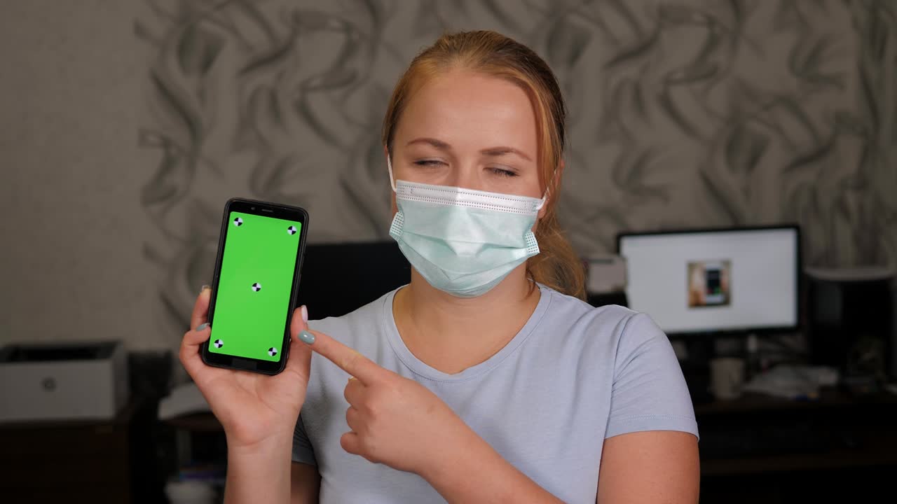 una mujer con una máscara médica sostiene un teléfono con una pantalla verde en casa.