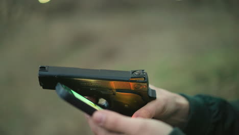 una vista de cerca de un hombre con una chaqueta verde sentado, sosteniendo un arma en una mano y un teléfono celular en la otra operando el teléfono celular con su pulgar, mientras la luz se refleja en el arma