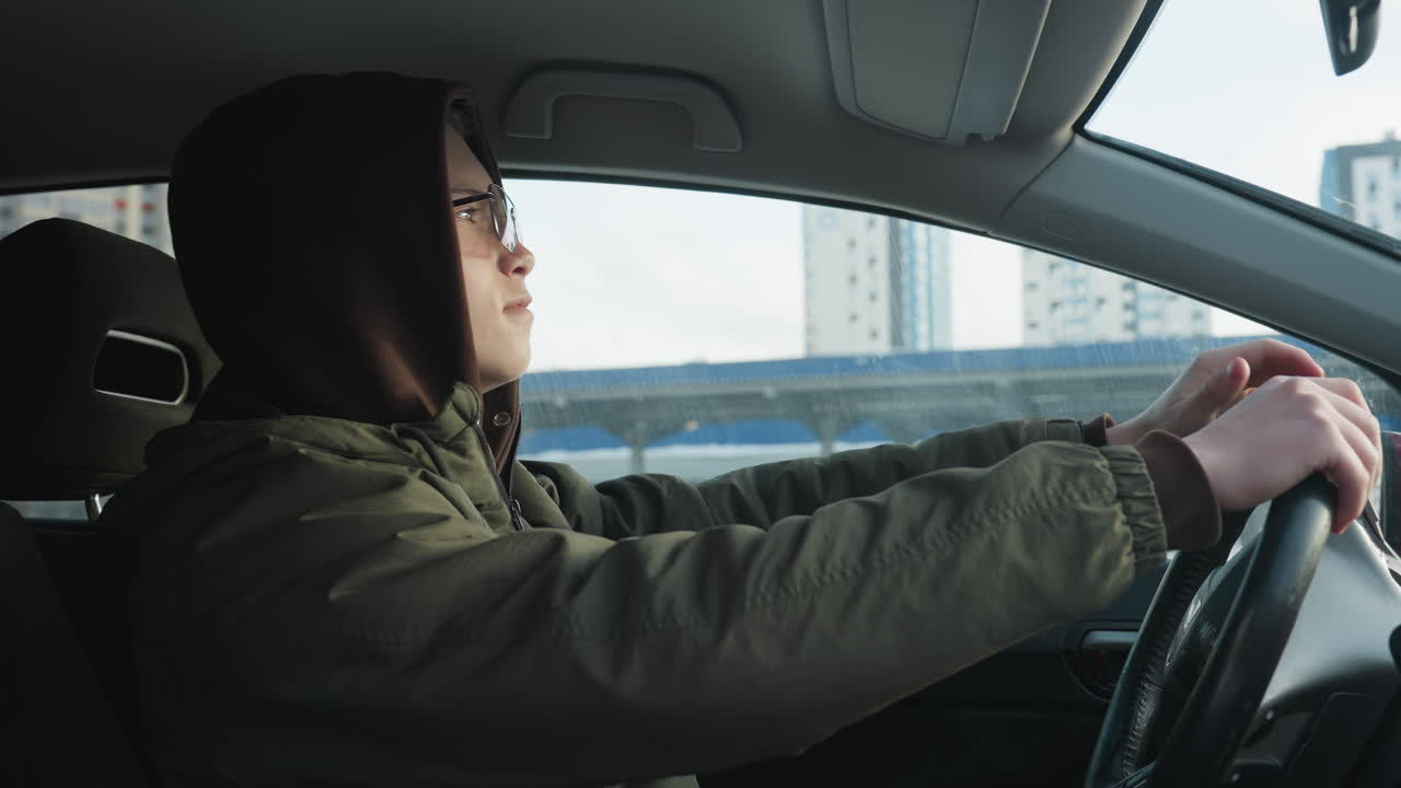 young man in hoodie sits in parked car side view hands on steering wheel dancing to upbeat music through tinted window against urban winter backdrop sunlight glint on windshield relaxed energy bright