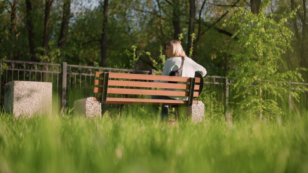 Blonďatý bělošský kytarista na lavičce hraje na akustickou kytaru v prosluněném jarním parku, uvolněný skladatel skládá melodii, ležérní džínová bunda, popředí s přírodní trávou, pozadí s alejemi, upřímný profil