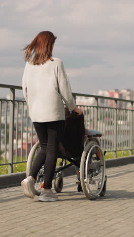 woman pushes wheelchair with child walking in modern park overlooking city panorama. mom on walk with little girl after surgery backside view