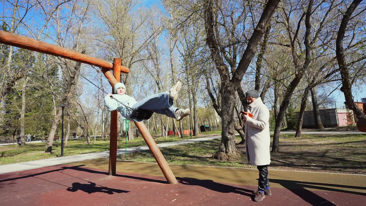 Niño columpiándose en el parque infantil mientras un adulto mira el teléfono en el parque