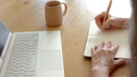 Mature-woman-writing-in-notebook-next-to-laptop-on-wooden-table