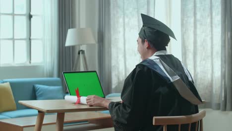 excited asian man showing off a university certificate to laptop computer with green mock-up screen during an online video call. male graduate wearing a graduation gown and cap sitting on the living room