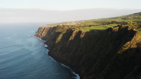 Drohnenaufnahmen-Einer-üppig-Grünen-Vulkaninsellandschaft-Mit-Hellem-Sonnenlicht-Und-Häusern-In-Der-Nähe-Von-Meeresklippen-Auf-Der-Azoreninsel-Sao-Miguel