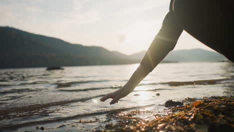 woman dips hand into lake water at sunset closeup. lady touches calm water enjoying beauty of nature in peaceful autumn evening. finding joy in nature