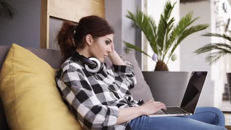 side view of a pretty girl in a plaid shirt and jeans relaxing on a couch with a laptop on legs and headphones on neck. room with a flower