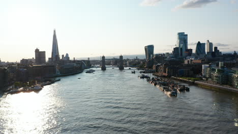 Landing-footage-above-Thames-river-against-sunshine.-silhouettes-of-Tower-Bridge-and-skyscrapers-on-river-banks.-London,-UK