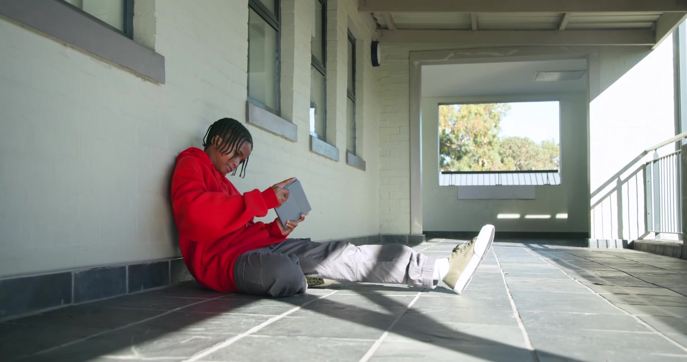 African American youth male sitting on tiled porch wearing red hoodie, tapping tablet, copy space