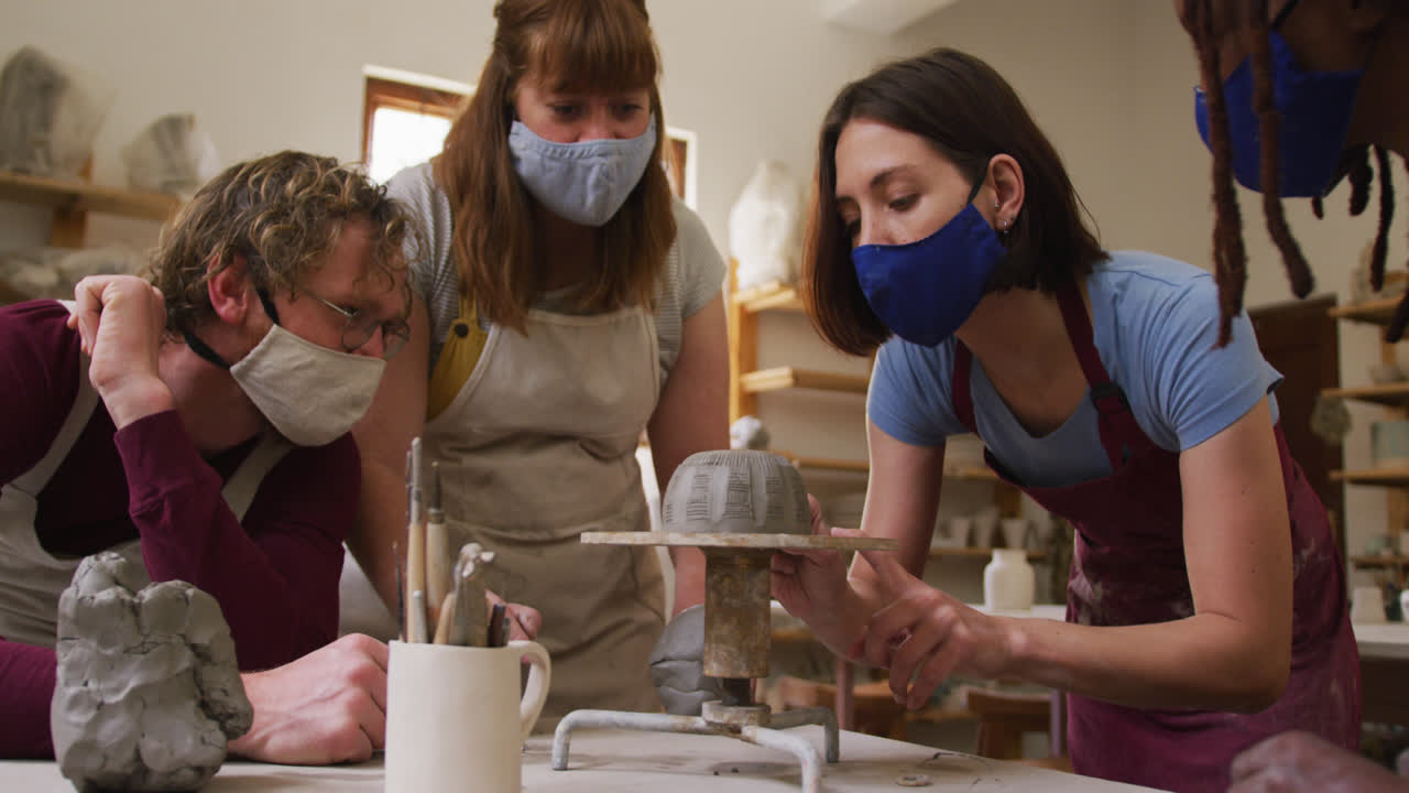Diverse potters wearing face masks and aprons working on pot on potters wheel at pottery studio