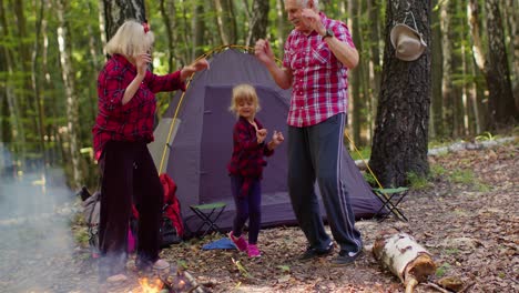 Senior-elderly-grandmother-grandfather-with-granddaughter-dancing-celebrating-over-campfire-in-wood.