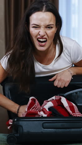 angry woman with suitcase indoors. young female trying to close the travel bag full of clothes in the hotel room. tourist girl packaging luggage. vertical video