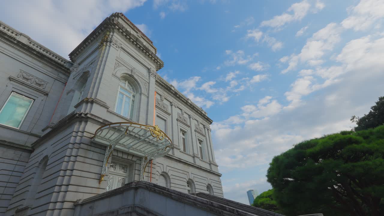 A striking low-angle shot of the majestic, stone side facade and portico of the Akasaka Palace against a beautiful cloudy blue sky