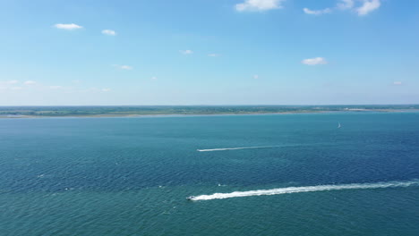 Aerial-shot-of-a-boat-moving-across-the-ocean-with-coastline-in-the-background,-on-a-bright-sunny-day