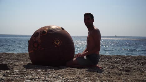 hombre haciendo ejercicio de yoga al aire libre cerca de una vieja mina marina flotante oxidada en la playa con costa rocosa y fondo marino. estilo de vida saludable, contaminación, protección de la naturaleza, concepto de guerra y paz