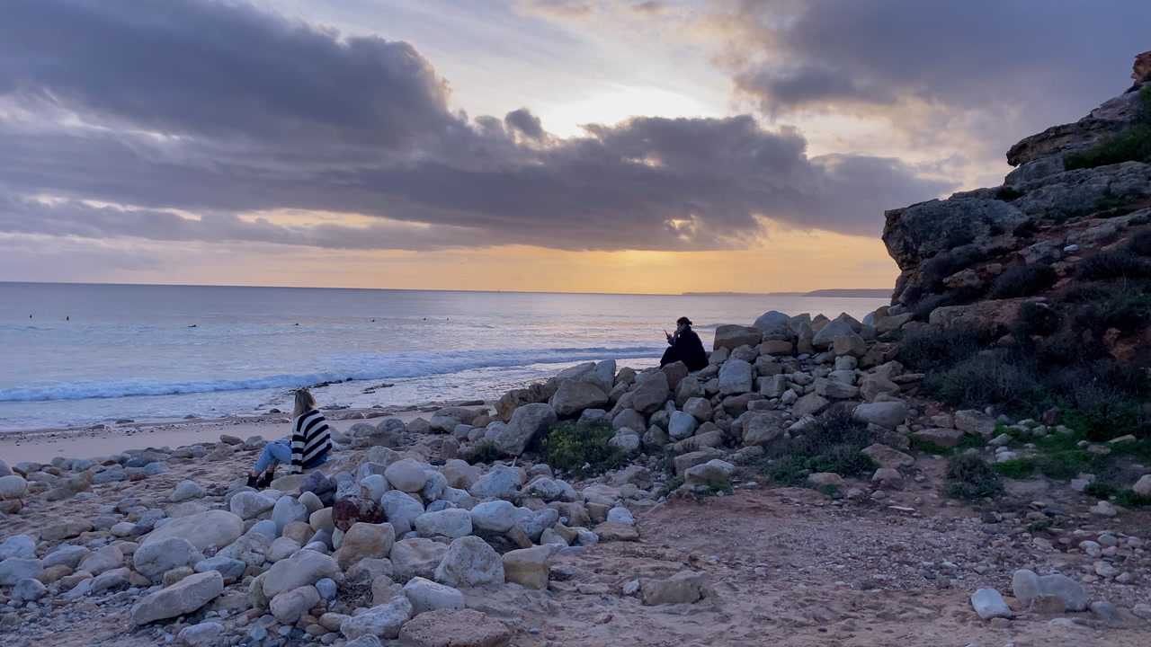 Two people enjoying a dramatic sunset on a rocky beach, watching waves and a few surfers in the distance