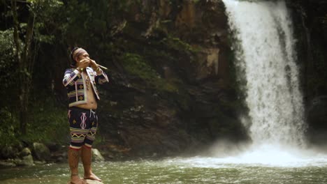 young man playing bamboo flute behind is a waterfall while standing