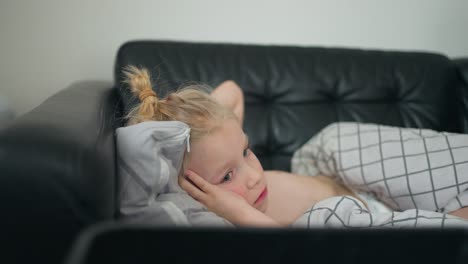 in a close-up head shot, a 7-year-old caucasian boy lies on a black couch, finding solace in watching tv during moments of nausea