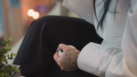 close up of tattooed hand squeezing stress ball against black trousers, with soft indoor lighting and blurred plant in foreground, capturing calm and focused emotional release moment