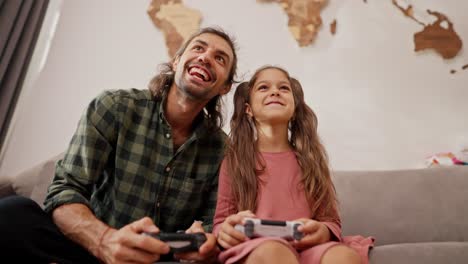 A-little-brunette-girl-with-a-ponytail-hairstyle-in-a-pink-dress-plays-with-a-joystick-together-with-her-happy-father,-a-brunette-man-in-a-Green-checkered-shirt-sitting-on-a-gray-sofa-in-a-modern-apartment