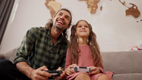 una pequeña chica morena con un peinado de cola de caballo en un vestido rosa juega con un joystick junto con su padre feliz, un hombre moreno en una camisa a cuadros verdes sentado en un sofá gris en un apartamento moderno